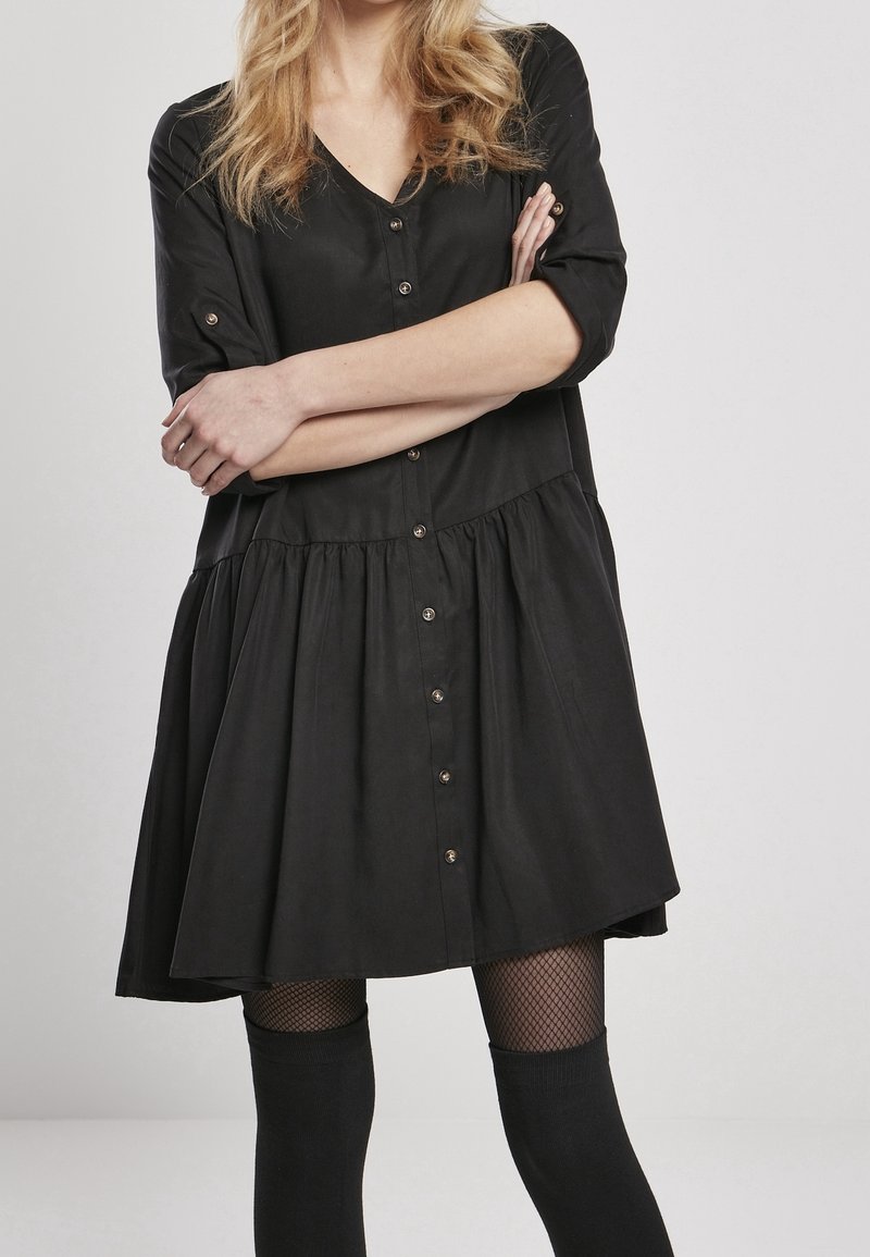 Robe noire boutonnée avec un col en V, des manches courtes et une jupe froncée. Elle est dotée de boutons métalliques et est fabriquée dans un tissu lisse.