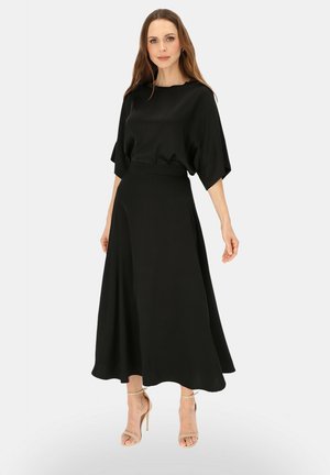 Kobieta o długich brązowych włosach, ubrana w czarną bluzkę z krótkim rękawem i zwiewną czarną spódnicę midi oraz beżowe sandały na obcasie na białym tle.