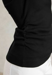 Čierny rebrovaný top s dlhým rukávom a detailným zberaním na boku, s priliehavým strihom a hladkou textúrou. V páre s bielou látkou, ktorá zvýrazňuje kontrast.