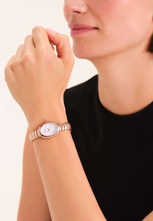 Orologio in oro rosa con quadrante bianco rotondo, cinturino a maglie e design minimalista, indossato al polso, visto di lato, su uno sfondo neutro.