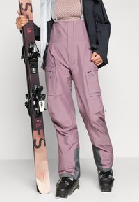 Lyžiarske nohavice svetlofialovej farby so sivými akcentmi, nepremokavý materiál, nastaviteľné traky a niekoľko vrecká na zips; kombinované s lyžami a lyžiarskymi topánkami.