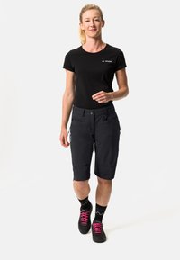 Schwarzes T-Shirt mit VAUDE-Logo, kombiniert mit knielangen dunklen Shorts mit Reißverschlusstaschen und schwarzen Schuhen mit pinken Schnürsenkeln.