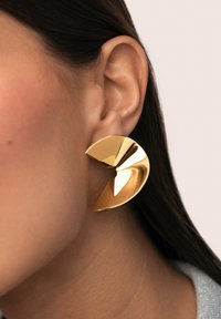 Pendientes de metal tono dorado con un diseño curvo y abstracto de textura pulida, que envuelven el lóbulo de la oreja para una apariencia llamativa.