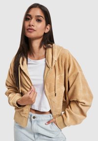 Sweat à capuche en velours côtelé de couleur beige clair, avec une surface texturée. Il présente une capuche avec cordon de serrage et des poches latérales, associé à un haut blanc.