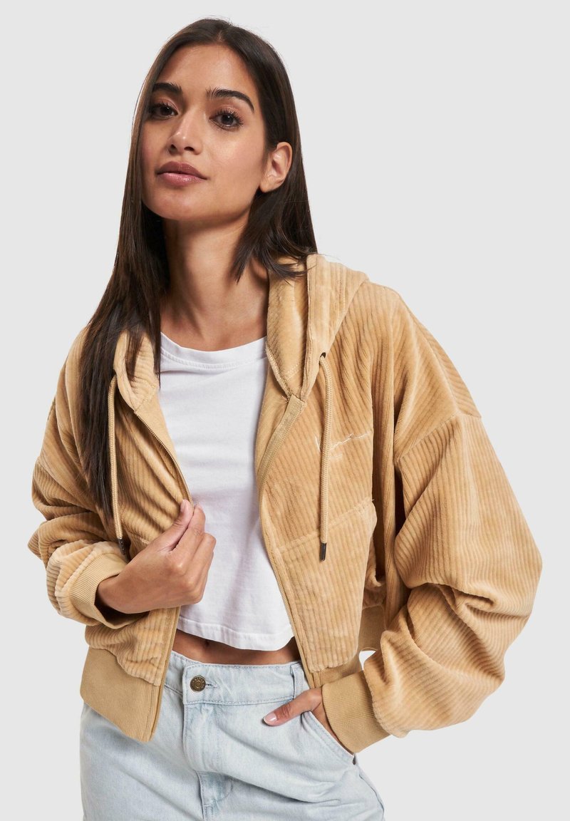 Sweat à capuche en velours côtelé de couleur beige clair, avec une surface texturée. Il présente une capuche avec cordon de serrage et des poches latérales, associé à un haut blanc.