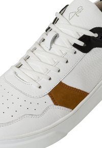Zapatilla blanca de cuero con un acento de gamuza marrón, cordones planos, caja de dedos perforada y detalles en negro cerca del área de los cordones.
