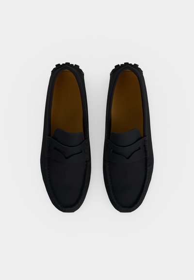 Ζευγάρι μαύρα δερμάτινα μοκασίνια με καφέ εσωτερική επένδυση και λάστιχα στη σόλα, εμφανίζονται από πάνω σε λευκό φόντο.