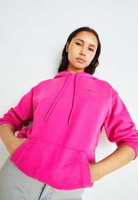 Felpa rosa con cordini, tasca a marsupio e polsini a costine. Presenta un piccolo tag con logo sul petto. Il materiale sembra morbido e strutturato.