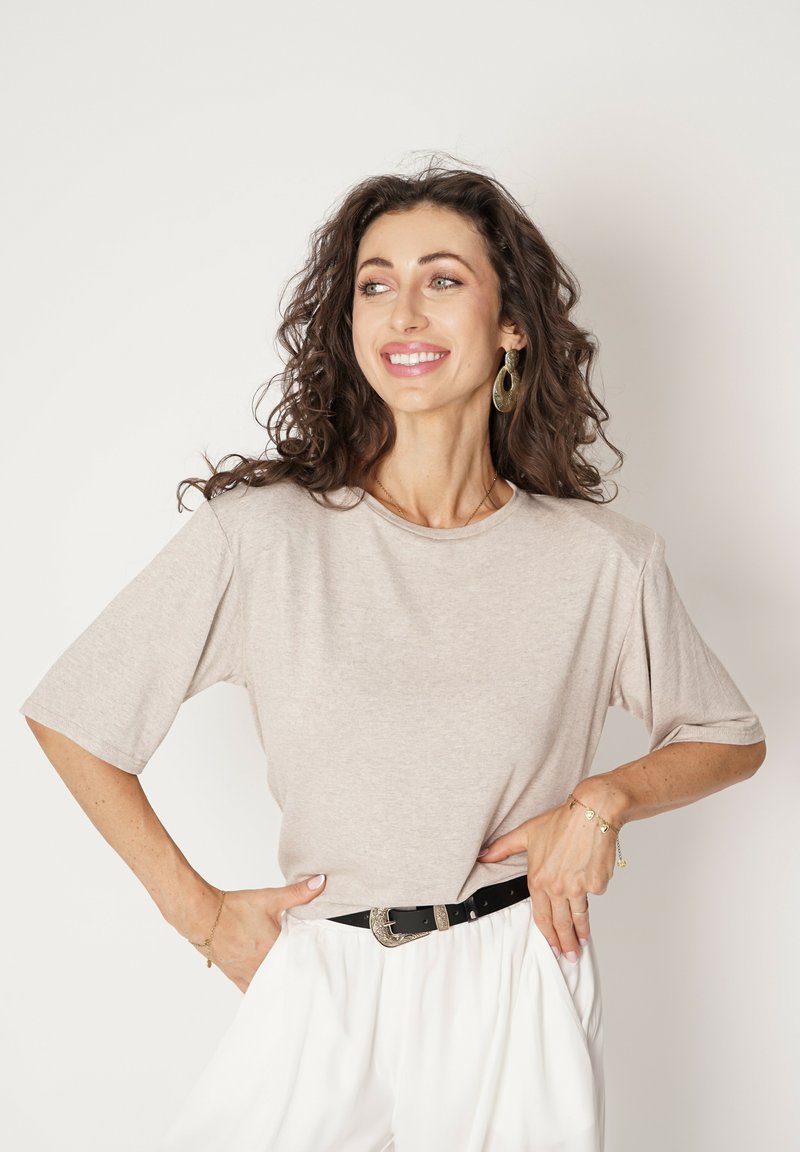 Beżowa koszulka z krótkim rękawem o luźnym kroju, noszona z czarnym paskiem. Kręcone włosy modelki oprawiają jej twarz na białym tle.