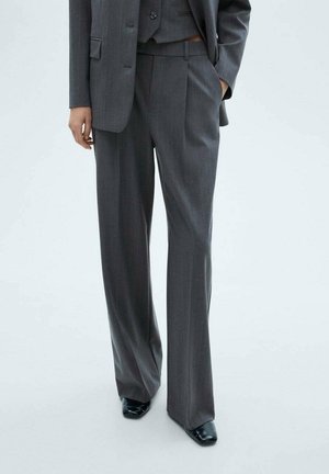 Osoba nosząca ciemnoszare szerokie spodnie w drobną prążek, pasującą marynarkę oraz czarne buty z kwadratowym noskiem, stojąca z jedną ręką w kieszeni.