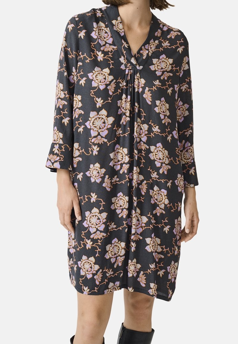 Femme portant une robe noire mi-longue avec des motifs floraux roses et beiges et des manches trois-quarts, associée à des bottes noires.