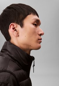 Čierna prešívaná vesta s vysokým golierom, vybavená zipsom a šnúrkou, nosená cez tmavú košeľu, z profilu na sivom pozadí.