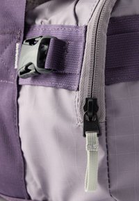 Κοντινό πλάνο τμήματος μωβ υφασμάτινης τσάντας με μαύρο τράβηγμα φερμουάρ και πλαστική πόρπη πλευρικής απελευθέρωσης σε λουρί που ταιριάζει.