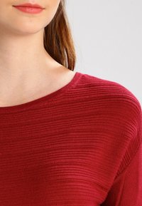 Haut en tricot rouge présentant un col rond et un tissu texturé avec des rayures horizontales. Les manches sont légèrement tombantes pour une coupe décontractée.