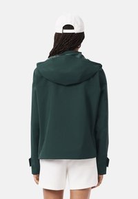 Persoon met vlechten draagt een donkergroene hoodie, witte shorts en een witte baseballcap, met de rug naar de camera tegen een effen achtergrond.