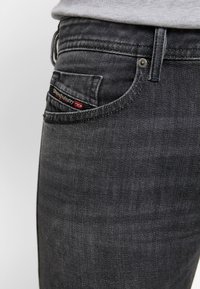 Dark grey denim jeans with a fitted design. Features include a back pocket with a red and black logo tag, and subtle fading detail.