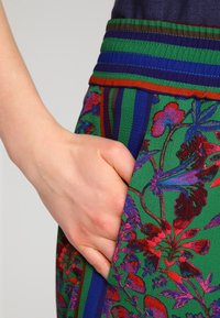 Tissu vert à motif floral avec des rayures rouges et bleues, doté d'une poche latérale et d'une finition texturée. Gros plan sur une main reposant dans la poche.