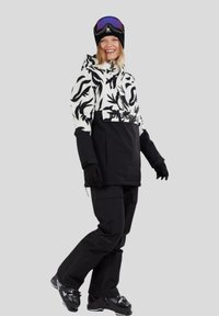 Donna sorridente con giacca da sci a motivi bianco e nero, pantaloni neri, guanti, stivali da snowboard e occhiali da sci viola su un berretto nero.