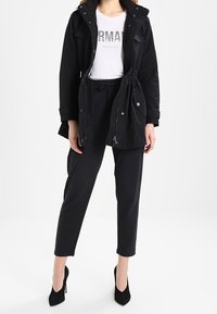 Chaqueta parka negra con capucha, bolsillos frontales y cintura con cordón, llevada sobre una camiseta gráfica blanca y pantalones negros ajustados. Tacones negros.