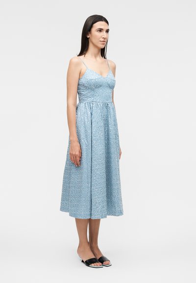 Γυναίκα με ίσια σκούρα μαλλιά που φοράει ένα αμάνικο, ανοιχτό μπλε φόρεμα με σχέδια μέχρι τη μέση της γάμπας και μαύρα ανοιχτά φλατ σανδάλια, στέκεται πλάγια.