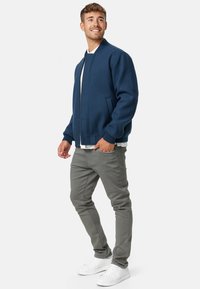 Marineblauwe bomberjack van stof met een gladde textuur, voorzien van ribgebreide manchetten en zoom, gecombineerd met lichtgrijze broek en witte sneakers.