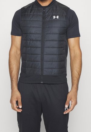 Mężczyzna noszący czarną pikowaną bezrękawnik na czarnej koszulce z krótkim rękawem i czarnych spodniach, z widocznymi logo Under Armour.