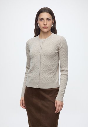 Kvinde iført en beige kabelstrikket cardigan med perleknapper og et brunt skørt, stående mod en ensfarvet hvid baggrund.