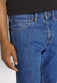 Gros plan d'une personne portant un jean en denim bleu avec un petit logo rectangulaire près de la poche avant et un t-shirt noir.