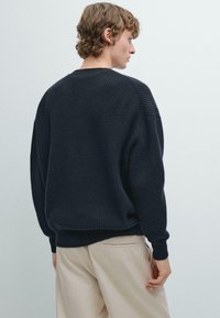 Mörk marinblå tröja med ribbad textur, rund halsringning och sänkta axlar. Matchas med beige byxor, subtil stickad detalj på ärmarna.