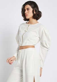 Weißes, tailliertes Jacket mit Reißverschluss, puffierten Ärmeln und glatter Textur, kombiniert mit hoch taillierten weißen Hosen mit seitlichem Schlitz.