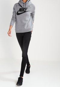 Felpa grigia Nike con un grande logo nero, abbinata a jeans skinny neri e scarpe sportive nere, che cammina su uno sfondo neutro.