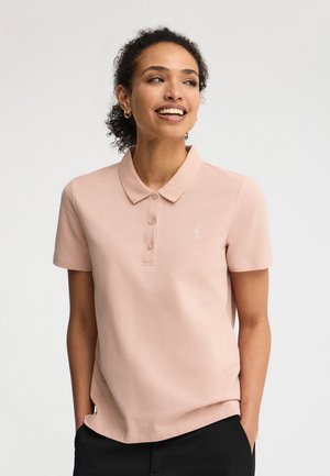 Kobieta uśmiechająca się, ubrana w jasnoróżową koszulkę polo z krótkim rękawem z małym białym logo i czarne spodnie, ręce w kieszeniach, na jednolitym tle.