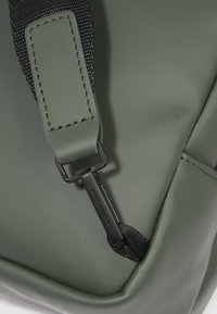 Dettaglio di una borsa verde in poliuretano con tracolla nera cucita, accessori metallici di fissaggio e superficie testurizzata.