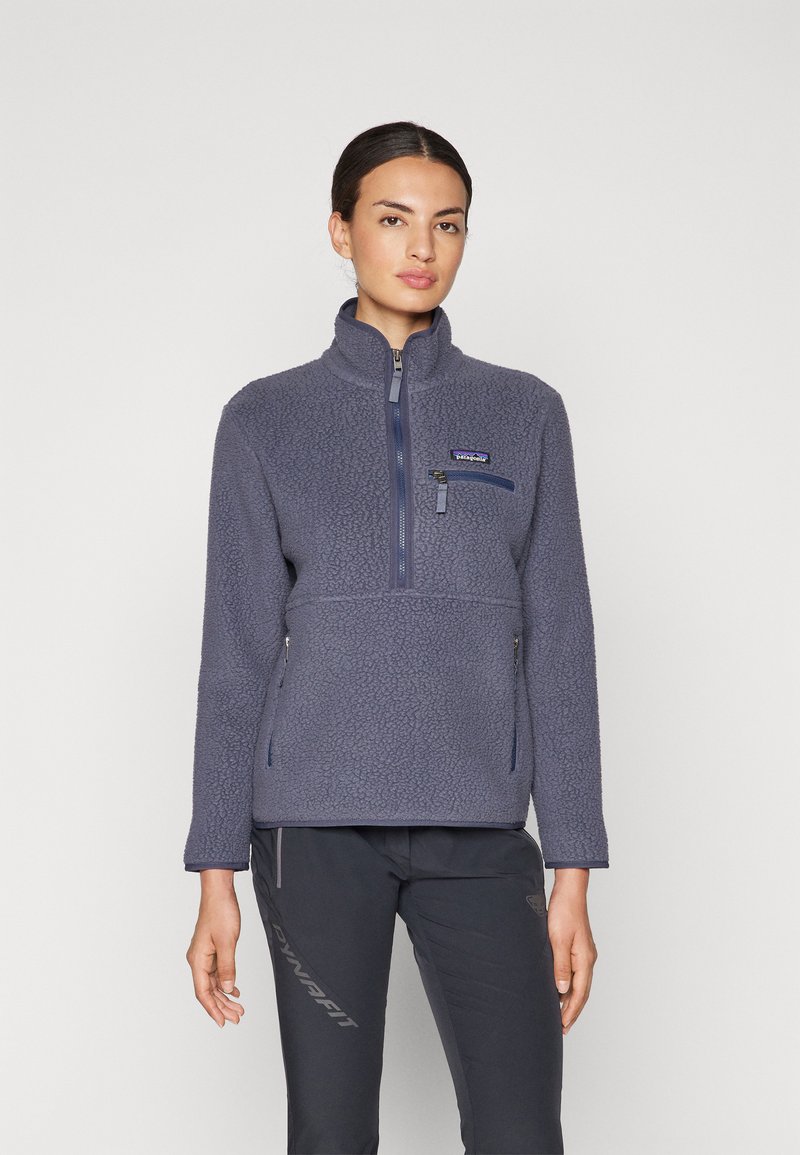 Felpa in pile di colore blu scuro con colletto alto, chiusura frontale a metà zip e due tasche laterali con zip; tessuto morbido e testurizzato.