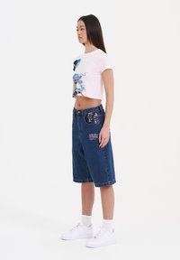 Camiseta corta rosa con gráfico de dragón azul, combinada con pantalones cortos de mezclilla oscuros tipo Bermuda que presentan acentos florales y de logotipo. Zapatillas blancas.
