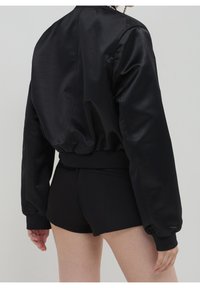 Veste bomber en satin noir avec une silhouette décontractée, une taille côtelée et des manches froncées. Associée à un short noir ajusté.