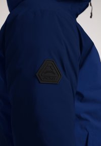 Mørkeblå jakke med et struktureret sort hexagonalt mærke med ordet "JACKIT" og geometriske linjer prægede på skulderen.