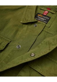 Chaqueta verde oliva con textura de algodón, que cuenta con un cuello, botones a presión y una etiqueta cerca del cuello con detalles de la marca e instrucciones de cuidado.