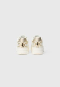 Scarpe da ginnastica bianche con accenti dorati metallici, dotate di pannelli posteriori texturizzati e un dettaglio logo prominente. Forma arrotondata e design con suola robusta.