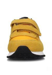 Sneaker gialla in suede con cinturini in velcro, suola in gomma nera e intersuola bianca. La vista frontale mette in mostra la forma arrotondata e la parte superiore testurizzata.