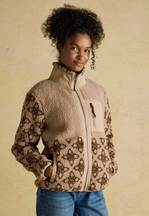 Woman with curly hair wearing beige patterned fleece jacket with zip and hands in pockets, standing against plain background.