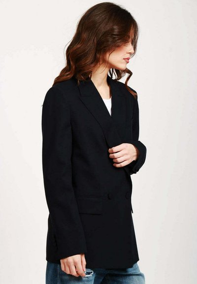 Blazer blu scuro realizzato in tessuto liscio, con un design doppiopetto, rever a scollo e due tasche frontali. Abbinato a dei jeans.