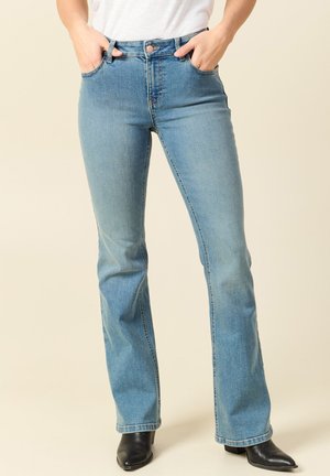 Osoba ubrana w jasnoniebieskie dżinsy z rozszerzanymi nogawkami, z rękami w kieszeniach i czarne botki na obcasie na beżowym tle.