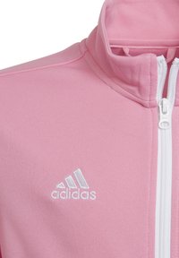 Rosa polyester zip-jacka med hög krage, vit dragkedja och en broderad vit Adidas-logga på vänster bröst.