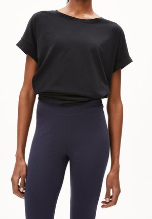 Kobieta ubrana w luźną czarną koszulkę z krótkim rękawem, wpuszczoną w wysokie, ciemnogranatowe legginsy, stojąca na jasnym, jednolitym tle.