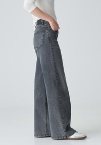 Persona con pantalones vaqueros de tiro alto y pierna ancha de color negro desgastado, zapatillas blancas y una camiseta de manga larga blanca, de pie con una mano en el bolsillo.