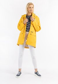 Chaqueta de lluvia amarilla impermeable con capucha, botones a presión, dos bolsillos frontales, combinada con leggings blancos y zapatillas azules. Camisa de rayas debajo.