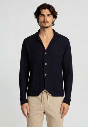Uomo che indossa un cardigan lavorato a maglia blu scuro con bottoni, due tasche frontali e pantaloni beige con coulisse, su sfondo neutro.