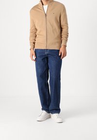 Beżowy sweter na zamek z prążkowanymi mankietami, zestawiony z niebieskimi jeansami o prostych nogawkach oraz białymi trampkami. Prosty design z minimalnym oznakowaniem marki.