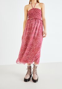 Kvinna iklädd en ärmlös rosa mönstrad midikjol som matchas med beige snörda kängor och vita strumpor mot en enfärgad bakgrund.